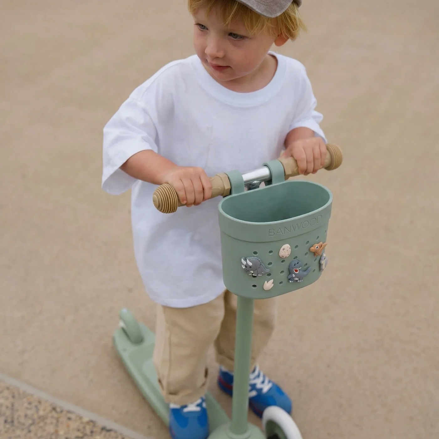 A young child wearing a cap and white shirt rides a green scooter with a front Banwood Kids’ Bike Basket on a paved surface, proudly displaying their collection of Banwood Kids’ Bike Basket Pins.