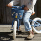 A child sits on a blue balance bike featuring Banwood’s Kids’ Bike Basket Pins, wearing rolled-up jeans, a light shirt, and brown boots, on a wooden surface outdoors.