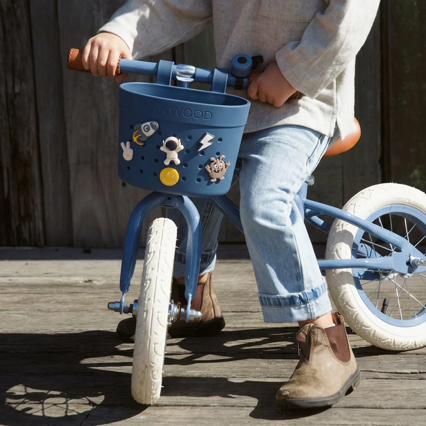 A child sits on a blue balance bike featuring Banwood’s Kids’ Bike Basket Pins, wearing rolled-up jeans, a light shirt, and brown boots, on a wooden surface outdoors.