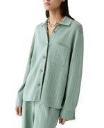 A woman wears the Pinstripe Work Shirt by Guest in Residence—a light green, pinstriped cotton-silk button-down—paired with matching Carpenter Pants and a chunky gold necklace.