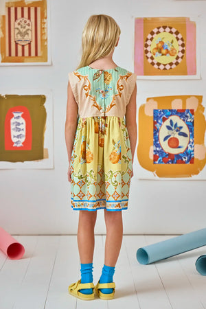 A young girl in the Girls' Piper Dress by Hunter Bell NYC stands on a white floor with blue socks and yellow sandals, facing wall-mounted art—her blonde hair shining in this charming kids' silhouette scene.