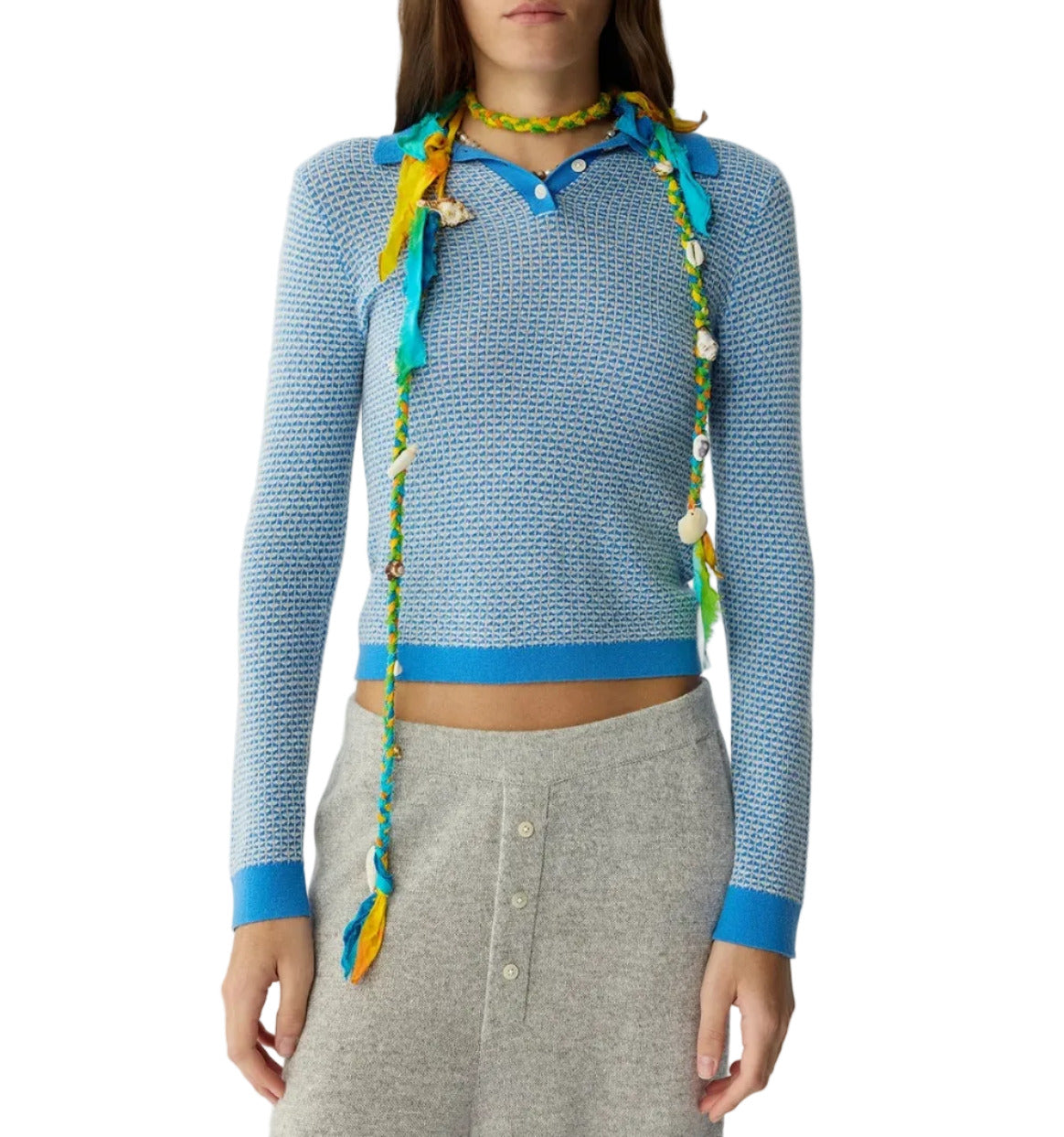 A person wears the Guest in Residence Pique LS Shrunken Polo in blue, paired with a light gray buttoned skirt and a vibrant necklace featuring beads, shells, and fabric accents.