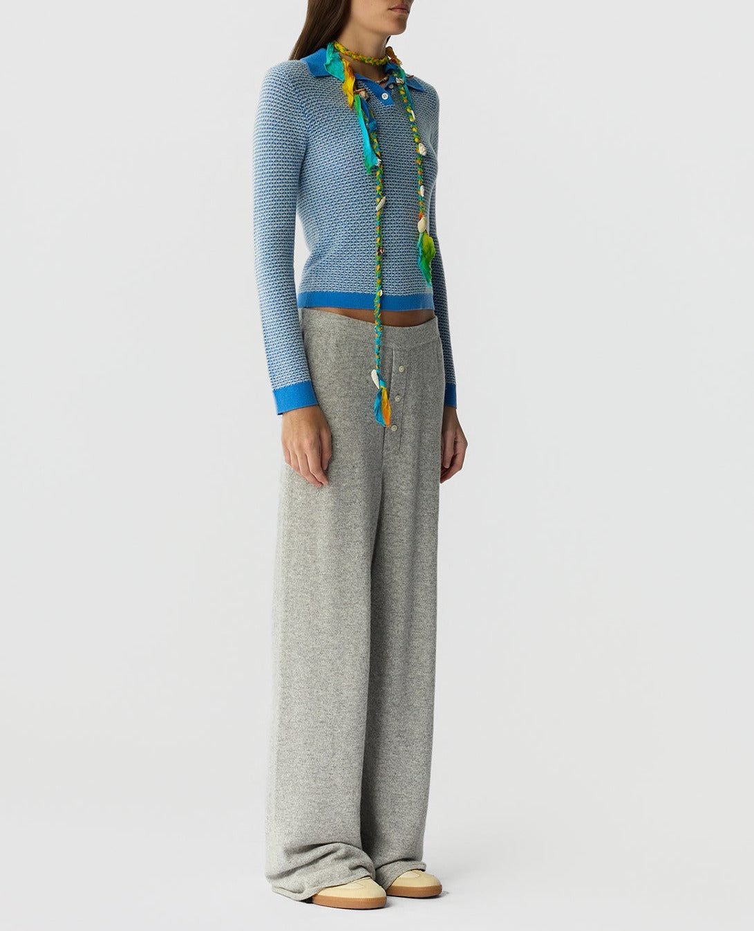 A person stands against a white background wearing the Guest in Residence Pique LS Shrunken Polo in light blue, styled with a colorful scarf, loose gray wide-leg pants, and beige shoes.