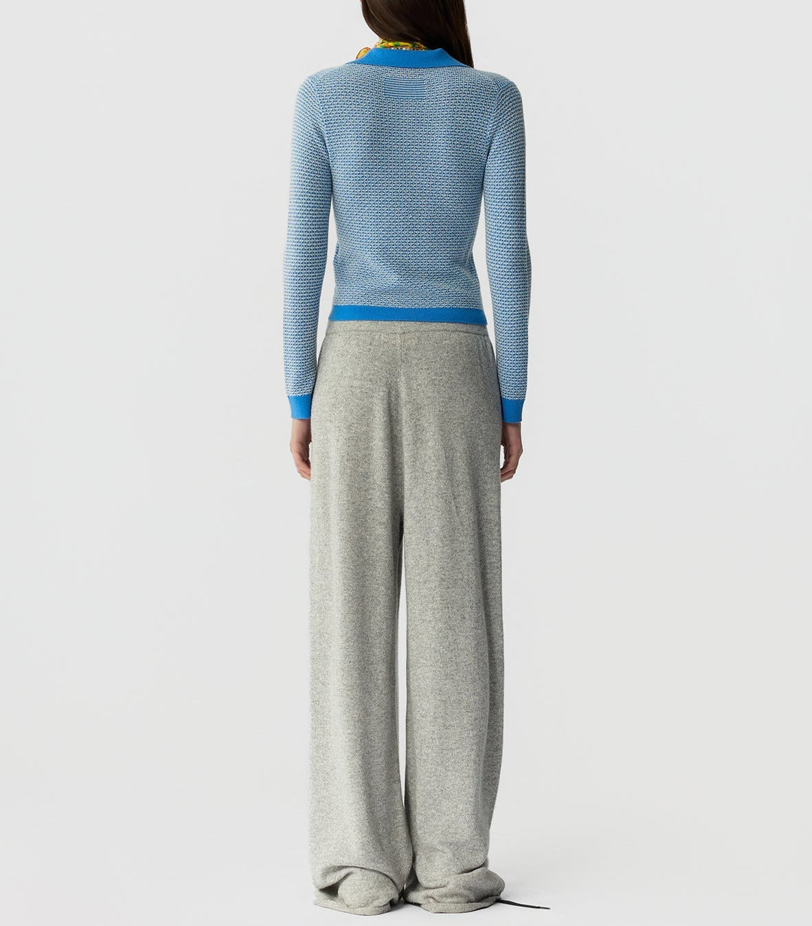 A person stands facing away in a light blue Guest in Residence Pique LS Shrunken Polo and loose-fitting gray pants, set against a plain white background.