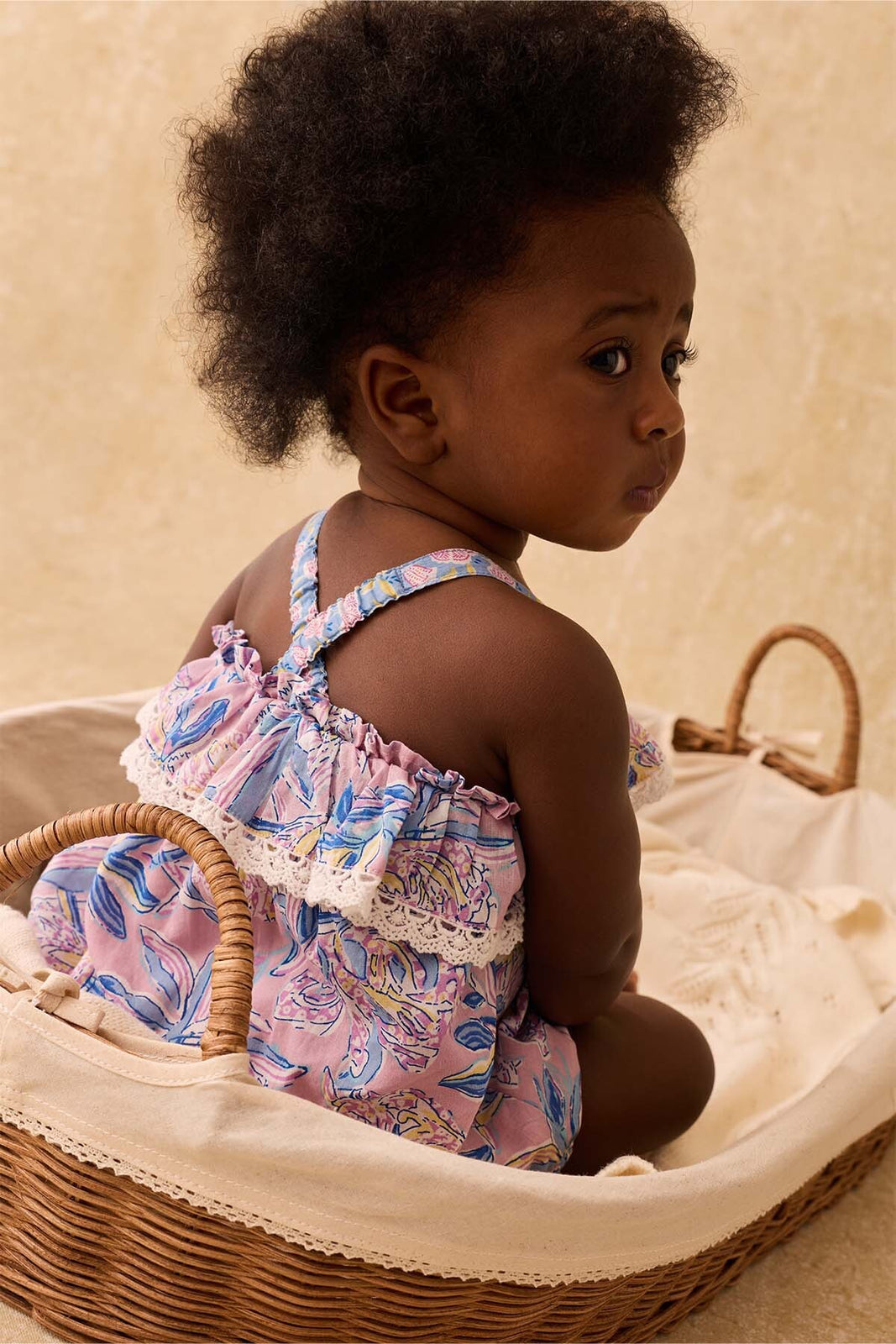 A young baby girl with curly hair sits in a wicker basket, wearing the Louise Misha Kumal Romper—a purple floral outfit with lace trim and a delicate cotton veil—looking off to the side against a beige background.