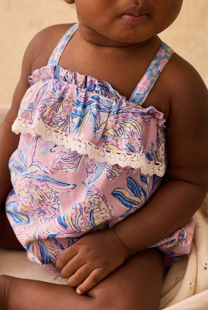 Baby girl wearing a pink floral romper with ruffles and lace trim, sitting down. Only the lower half of her face and body are visible.