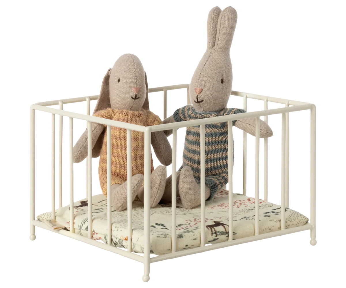 Two Maileg stuffed rabbits in knitwear sit inside a Maileg Playpen, Micro, placed on a patterned cushion.