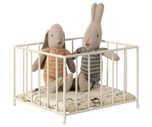 Two Maileg stuffed rabbits in knitwear sit inside a Maileg Playpen, Micro, placed on a patterned cushion.