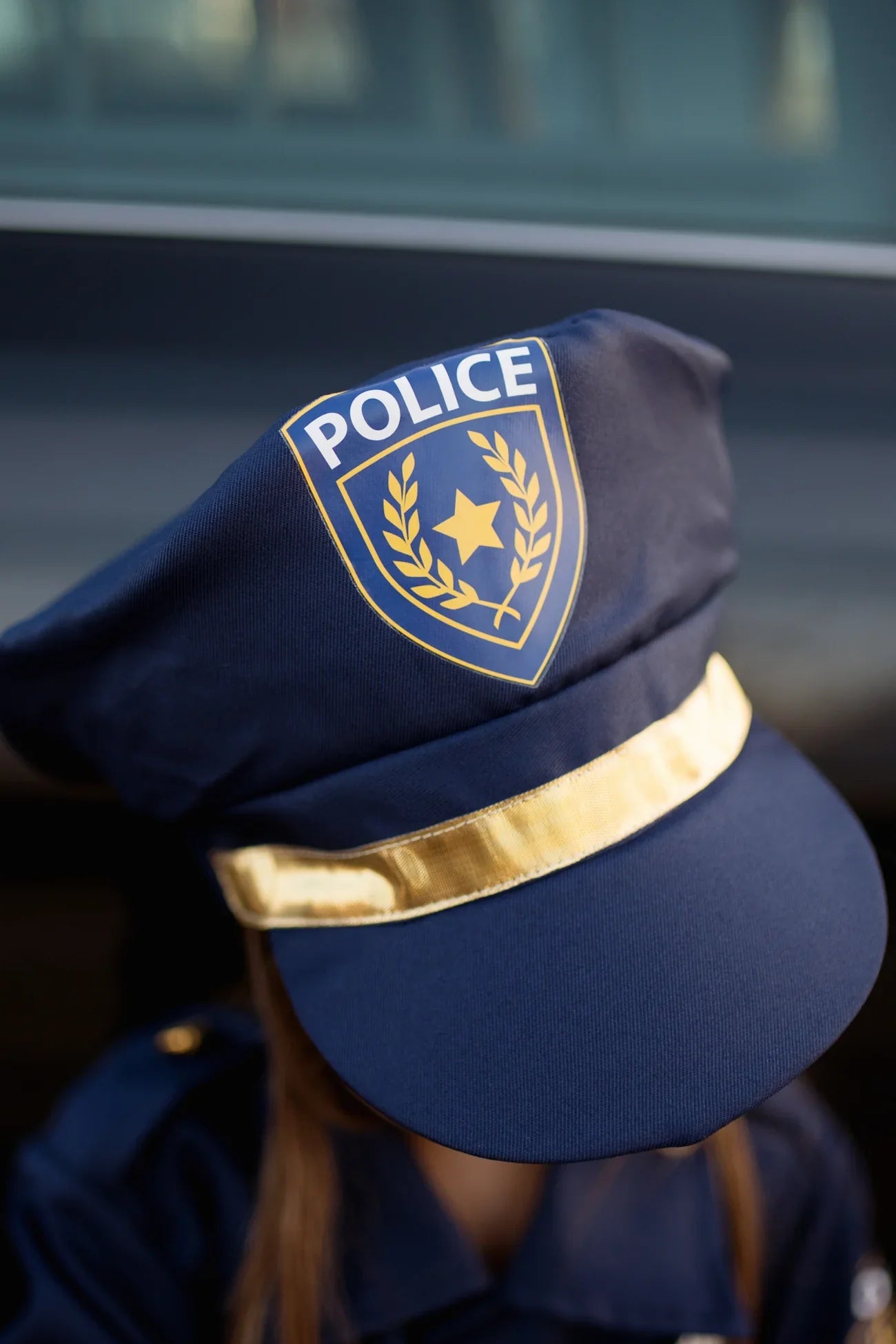 Someone wearing a navy blue police hat with gold band and badge from the Great Pretenders Police Officer Set; their face is mostly hidden, making it ideal for dress-up or imaginative play.