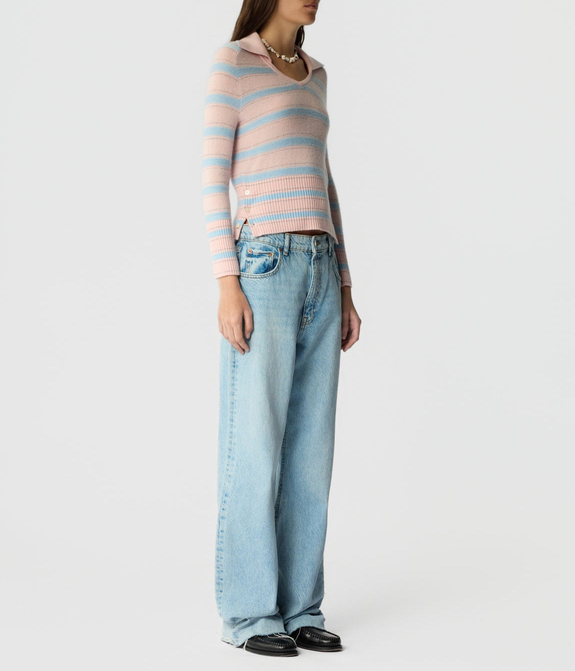 A person wears the Port Polo by Guest in Residence—pink and blue striped lightweight cashmere—paired with light blue baggy jeans and black shoes, standing against a plain white background.