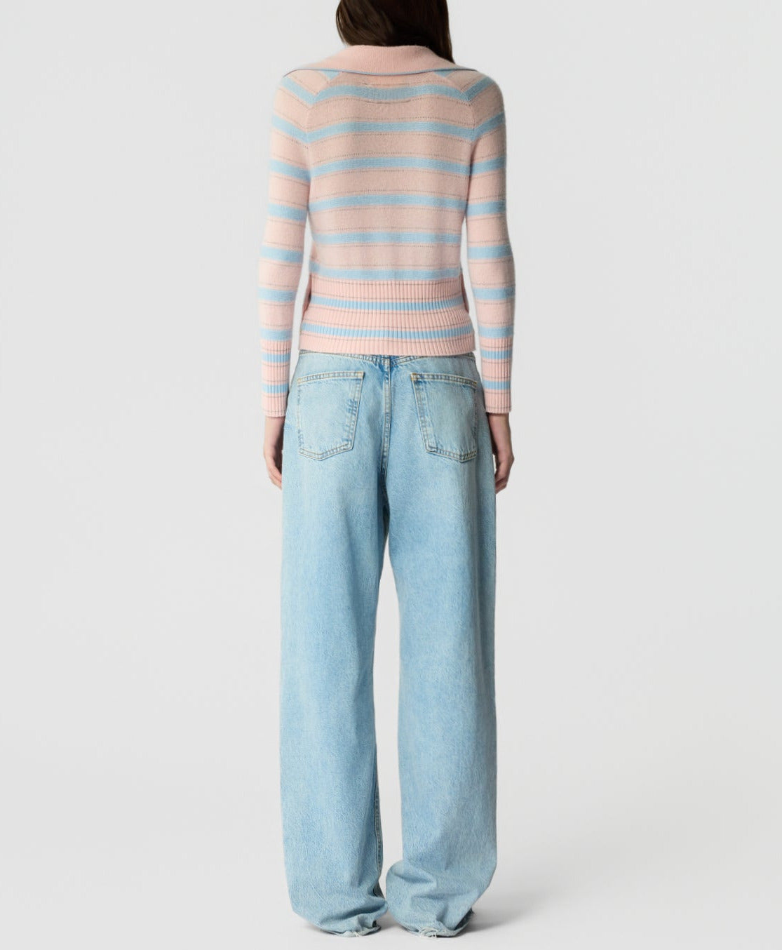 A person stands facing away, wearing the Port Polo in pastel pink and blue stripes by Guest in Residence, crafted from lightweight cashmere, paired with loose-fitting light blue jeans against a plain background.