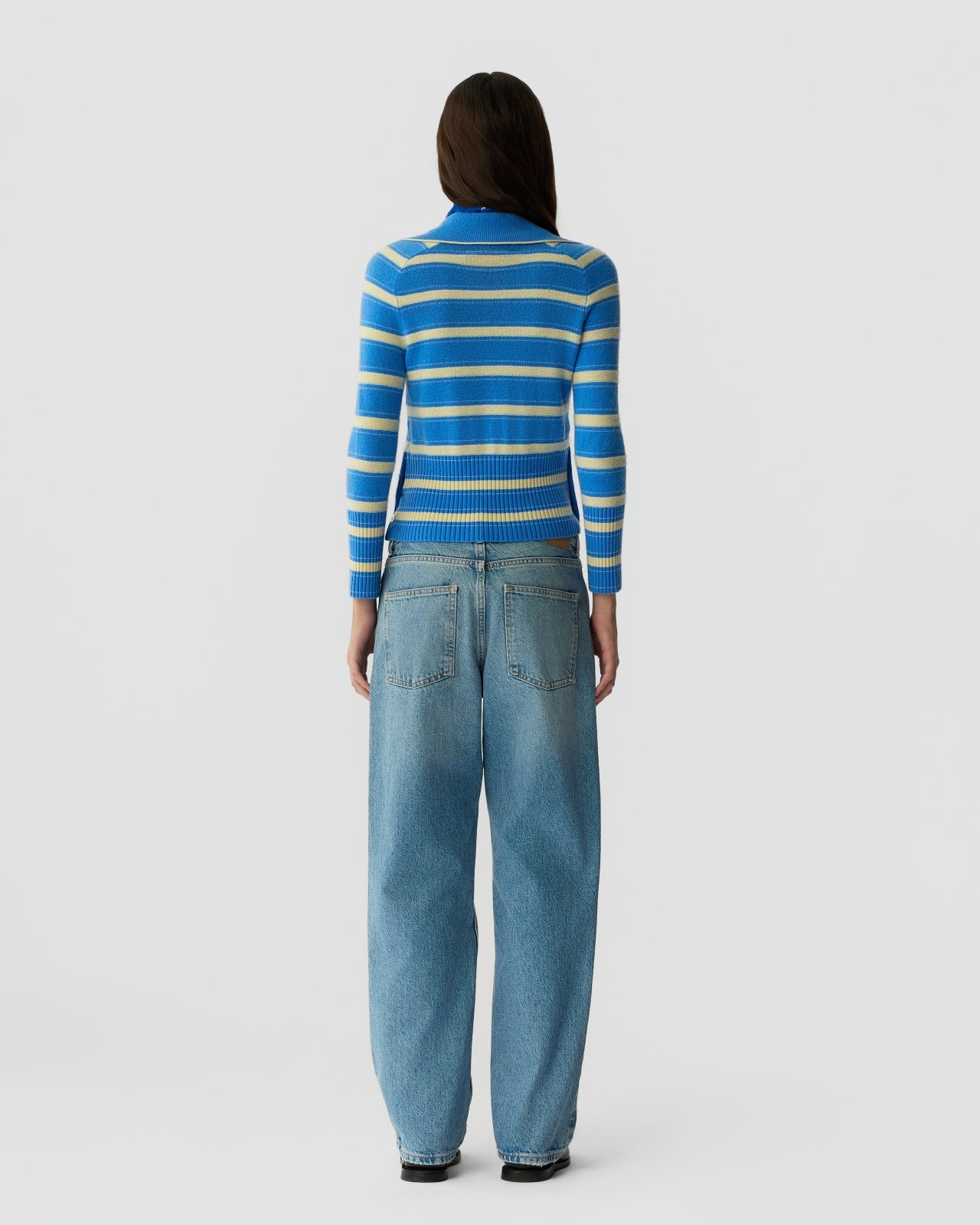 A person faces away, wearing the Port Polo by Guest in Residence—a blue and white striped sweater—paired with loose-fitting blue jeans against a plain light background.