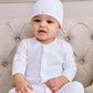 A baby, wearing the Kissy Kissy Premier Baby Duckling Hat with Hand Emb., and a white outfit, sits on a beige tufted couch facing the camera with a neutral expression.
