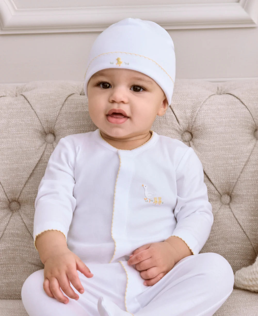 A baby, wearing the Kissy Kissy Premier Baby Duckling Hat with Hand Emb., and a white outfit, sits on a beige tufted couch facing the camera with a neutral expression.