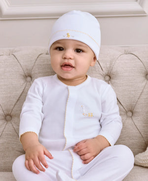 A baby in a white Kissy Kissy Baby Premier Baby Duckling Footie with Hand Embroidery sits on a beige tufted sofa, looking toward the camera.