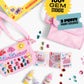 Two pink purses, gem stickers, paint bottles, and an instruction booklet from the Super Smalls Preppy Purse Gemify™ Craft Kit are arranged on a white background, showcasing a fun set for decorating your own stylish accessories.