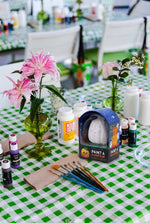 A table with a green checkered tablecloth is set with paint supplies, paintbrushes, vases of flowers, and a Paint a Ceramic Pumpkin Event kit from Hive for Kids—perfect for creating festive Halloween decorations after your Ceramic Pumpkin Painting Class in West Palm Beach.