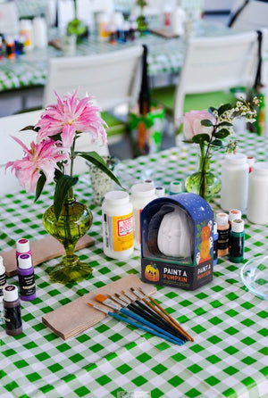 A table with a green checkered tablecloth is set with paint supplies, paintbrushes, vases of flowers, and a Paint a Ceramic Pumpkin Event kit from Hive for Kids—perfect for creating festive Halloween decorations after your Ceramic Pumpkin Painting Class in West Palm Beach.