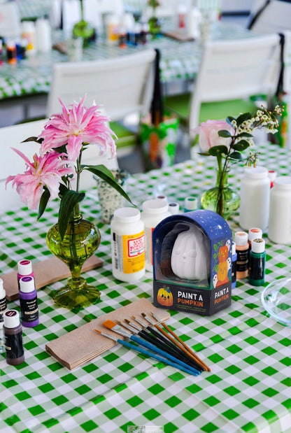 A table with a green checkered tablecloth is set with paint supplies, paintbrushes, vases of flowers, and a Paint a Ceramic Pumpkin Event kit from Hive for Kids—perfect for creating festive Halloween decorations after your Ceramic Pumpkin Painting Class in West Palm Beach.