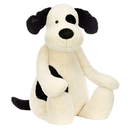 The Jellycat Bashful Black & Cream Puppy, Giant plush toy from Jellycat, showcases its endearing black ears and an adorable black spot encircling its right eye as it sits upright against a white backdrop. Filled with a cozy polyester inner filling, this soft toy is perfect for hugs and cuddles.