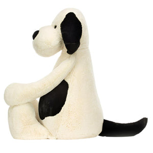 A side view of the Jellycat Bashful Black & Cream Puppy, Giant plush toy showcases its cream and black patches as it sits upright. This soft toy has a cozy polyester filling, offering cuddly comfort.