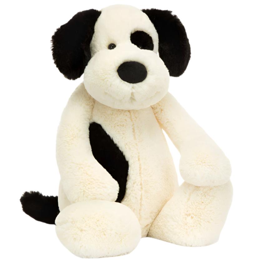 A really big Jellycat Bashful Black & Cream Puppy with a floppy ear and a patch over one eye sits upright, inviting cuddles.