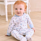A baby with red hair sits on a wooden floor, dressed in the Kissy Kissy Baby Boys’ Puppy Patch Zip Footie, made of soft Pima cotton with blue cuffs and a charming puppy patch print. A white chair can be seen in the background.
