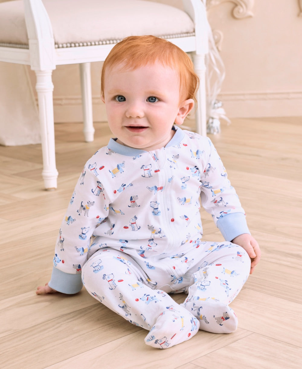 A baby with red hair sits on a wooden floor, dressed in the Kissy Kissy Baby Boys’ Puppy Patch Zip Footie, made of soft Pima cotton with blue cuffs and a charming puppy patch print. A white chair can be seen in the background.