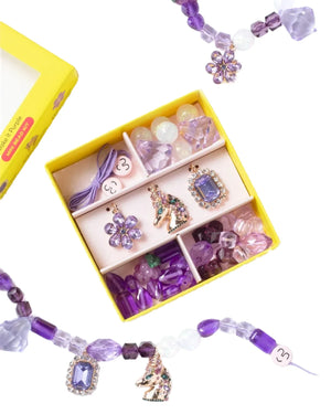 The Super Smalls Make It Purple Mini Bead Kit includes a yellow box with compartments that hold purple and pink beads, as well as special charms like a unicorn and a flower. A partially assembled beaded necklace wraps around, highlighting the appeal of unique jewelry from Super Smalls.