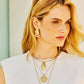 A woman with blonde hair wears the Jane Win Strong Along the Shore Earrings—bold gold statement pieces—paired with layered gold necklaces and a white top, standing in soft natural light.