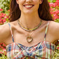 Woman wears a plaid dress and layered gold necklaces, including the Jane Win Love You to the Moon and Back Heart Pendant Necklace, smiling outdoors with flowers behind her.