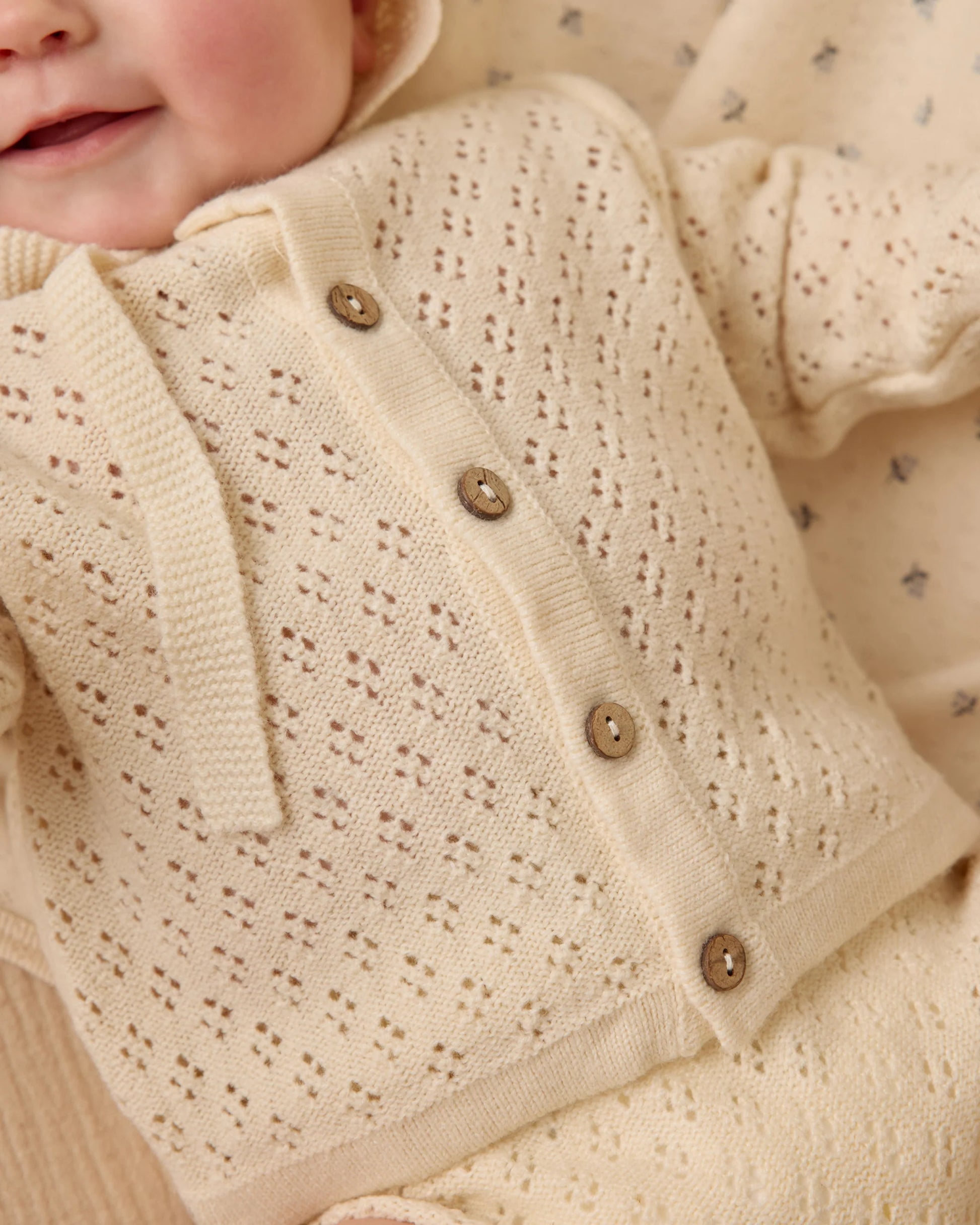 A baby in a cream Quincy Mae Baby Girls' and Girls' Knit Cardigan with coconut buttons and a matching outfit, lying on a patterned blanket.