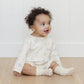 A baby with curly hair wears the Quincy Mae Baby Long Sleeve Romper, featuring a white printed design and paired with socks, while sitting on a wooden floor and smiling—ideal for everyday comfort.