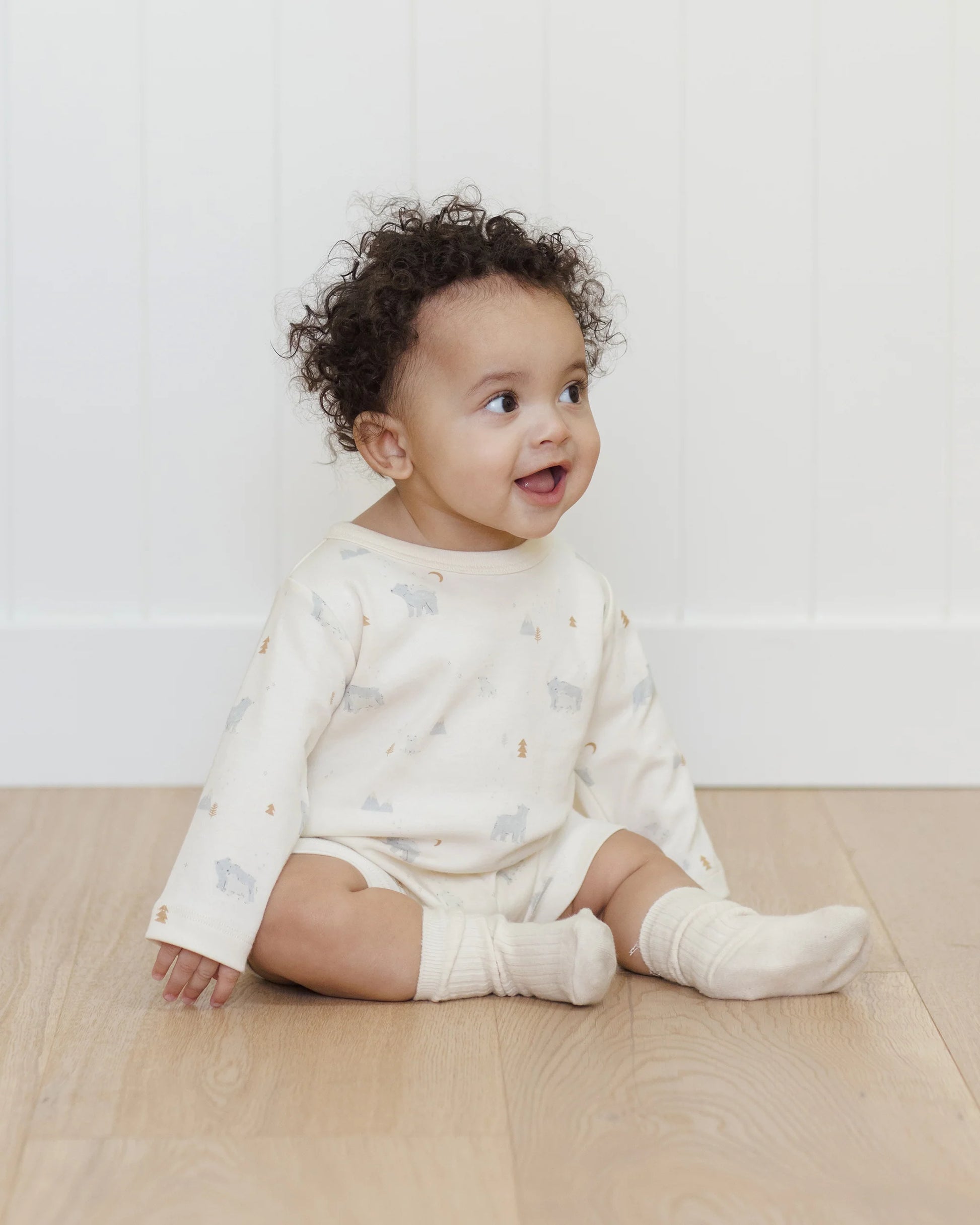 A baby with curly hair wears the Quincy Mae Baby Long Sleeve Romper, featuring a white printed design and paired with socks, while sitting on a wooden floor and smiling—ideal for everyday comfort.