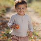 A young child stands outdoors holding an apple, dressed in the Quincy Mae Baby Boys’ and Boys’ Zion Shirt—an organic cotton, light checked shirt with a relaxed fit—and pinkish pants, against a softly blurred natural background.