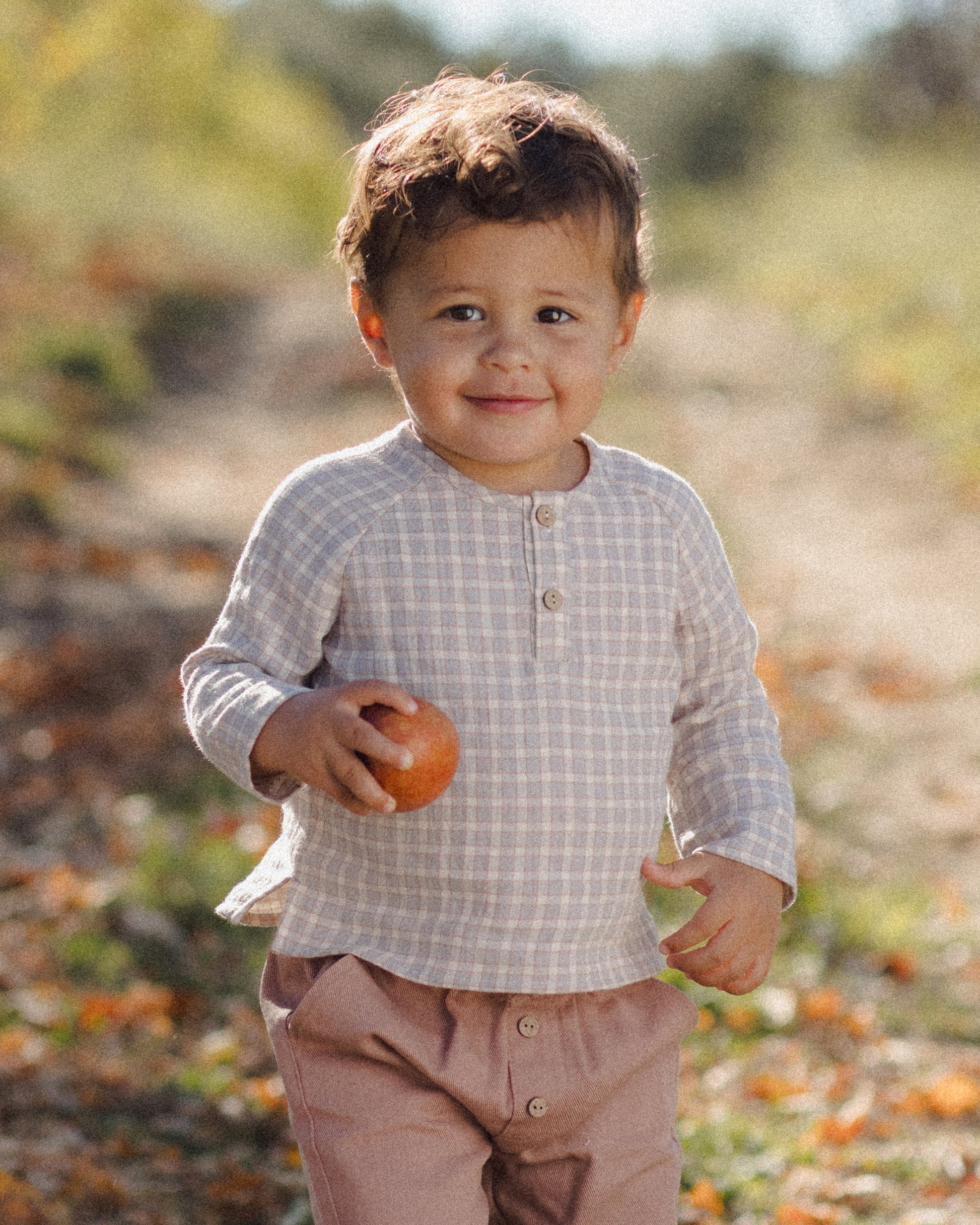 A young child stands outdoors holding an apple, dressed in the Quincy Mae Baby Boys’ and Boys’ Zion Shirt—an organic cotton, light checked shirt with a relaxed fit—and pinkish pants, against a softly blurred natural background.