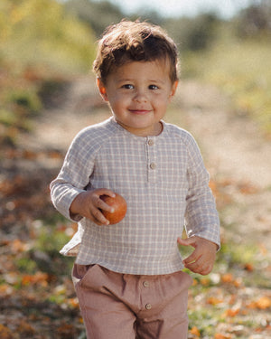 A young child stands outdoors holding an apple, dressed in the Quincy Mae Baby Boys’ and Boys’ Zion Shirt—an organic cotton, light checked shirt with a relaxed fit—and pinkish pants, against a softly blurred natural background.