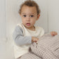 A young child with curly hair sits against a white wall, wearing a cream and gray shirt and Quincy Mae Baby Boys’ and Boys’ Woven Pant—relaxed fit, plaid with an elastic waistband—while holding a matching plaid fabric item.