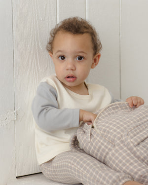 A young child with curly hair sits against a white wall, wearing a cream and gray shirt and Quincy Mae Baby Boys’ and Boys’ Woven Pant—relaxed fit, plaid with an elastic waistband—while holding a matching plaid fabric item.
