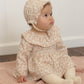 A baby wearing the Quincy Mae Baby Girls’ Thalia Romper with a matching bonnet sits on a pink textured blanket, looking to the side in front of a white wall.