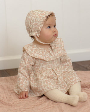 A baby wearing the Quincy Mae Baby Girls’ Thalia Romper with a matching bonnet sits on a pink textured blanket, looking to the side in front of a white wall.