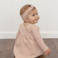 A baby wearing the Quincy Mae Baby Girl’s and Girls’ Ribbed Long Sleeve Dress in pink stripes, complete with coordinating bloomers, sits on a wooden floor facing slightly away from the camera, with a white wall in the background.