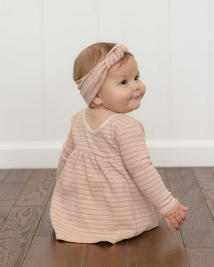 A baby wearing the Quincy Mae Baby Girl’s and Girls’ Ribbed Long Sleeve Dress in pink stripes, complete with coordinating bloomers, sits on a wooden floor facing slightly away from the camera, with a white wall in the background.
