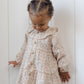 A young child with short curly hair stands against a white paneled wall, looking down while wearing the Quincy Mae Baby Girls’ and Girls’ Ruffle Collar Button Dress.