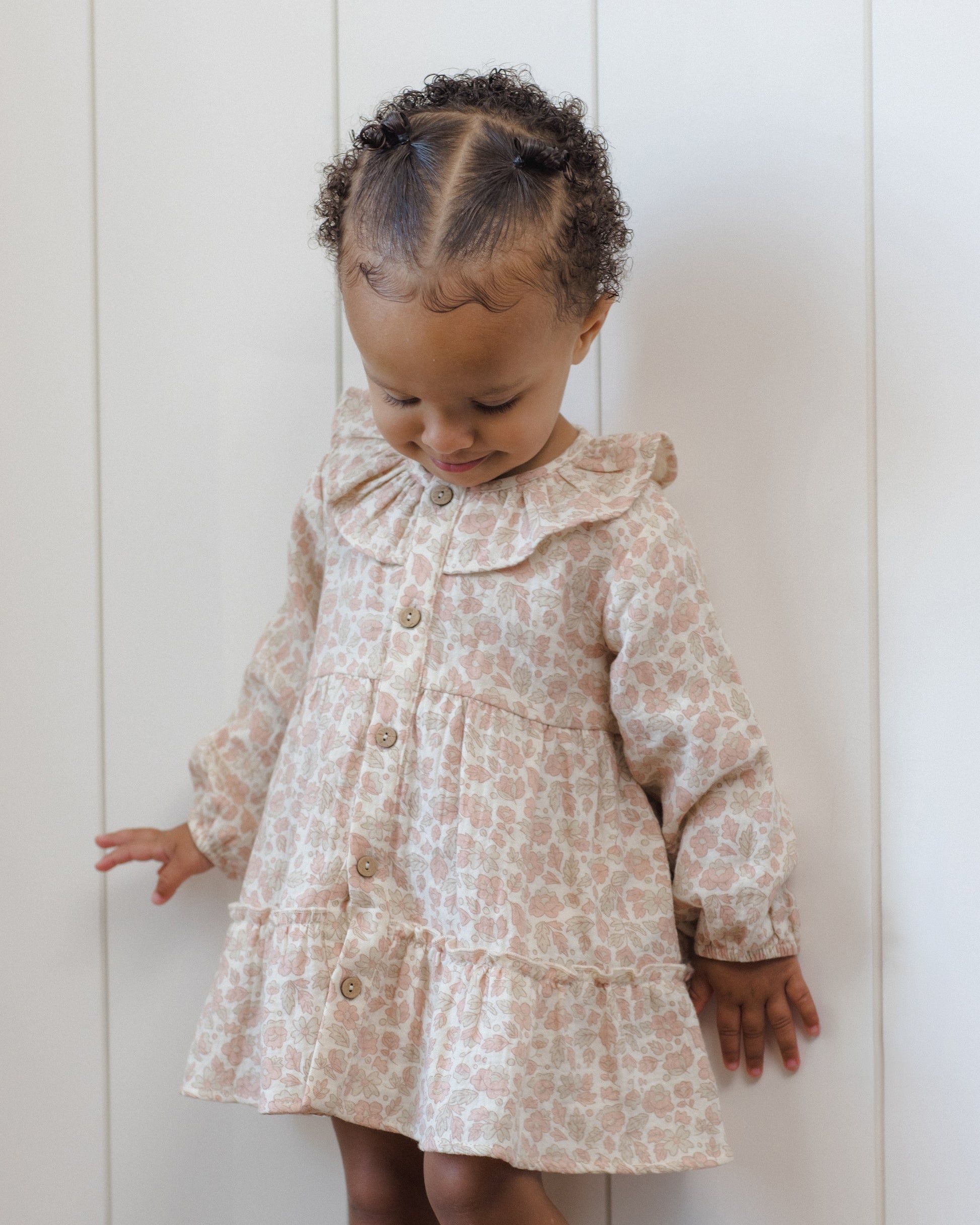 A young child with short curly hair stands against a white paneled wall, looking down while wearing the Quincy Mae Baby Girls’ and Girls’ Ruffle Collar Button Dress.