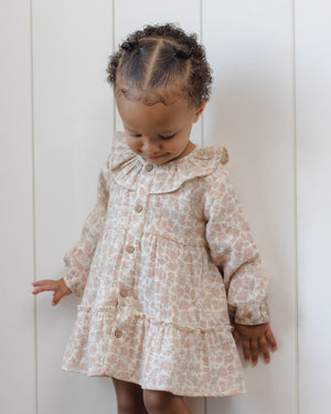 A young child with short curly hair stands against a white paneled wall, looking down while wearing the Quincy Mae Baby Girls’ and Girls’ Ruffle Collar Button Dress.
