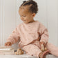 A young child wearing the Quincy Mae Baby Girls’ and Girls’ Relaxed Sweatshirt sits on a white ledge, holding a wooden toy car and touching a stack of books while looking to their left—perfectly embodying the essence of cozy essentials.