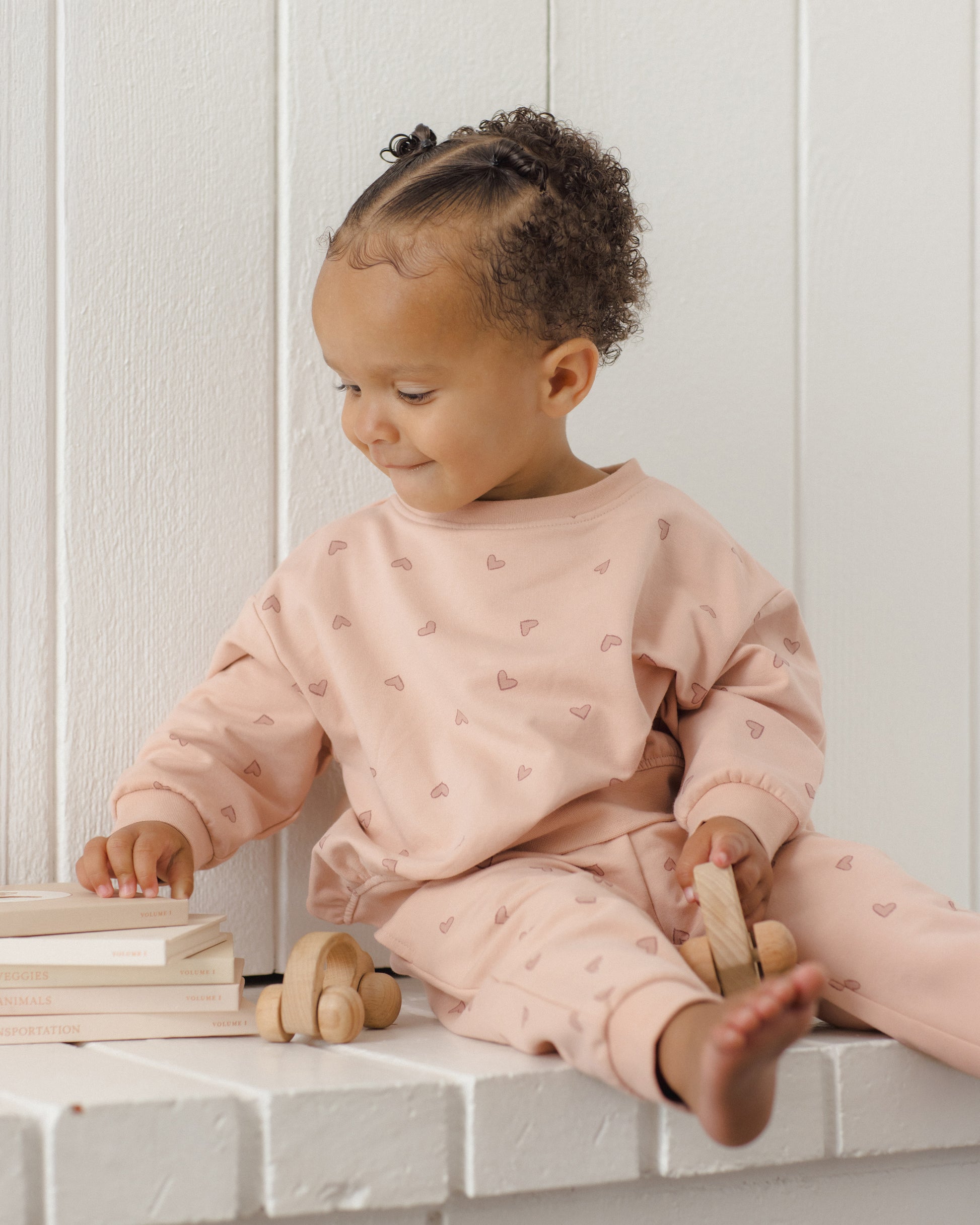 A young child wearing the Quincy Mae Baby Girls’ and Girls’ Relaxed Sweatshirt sits on a white ledge, holding a wooden toy car and touching a stack of books while looking to their left—perfectly embodying the essence of cozy essentials.