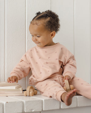 A young child wearing the Quincy Mae Baby Girls’ and Girls’ Relaxed Sweatshirt sits on a white ledge, holding a wooden toy car and touching a stack of books while looking to their left—perfectly embodying the essence of cozy essentials.