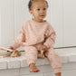 A toddler dressed in Quincy Mae’s Girls’ Relaxed Sweatpant, a cozy essential in pink covered with small heart patterns for all-day comfort, sits on a white brick ledge holding a wooden toy and gazing slightly to the side.